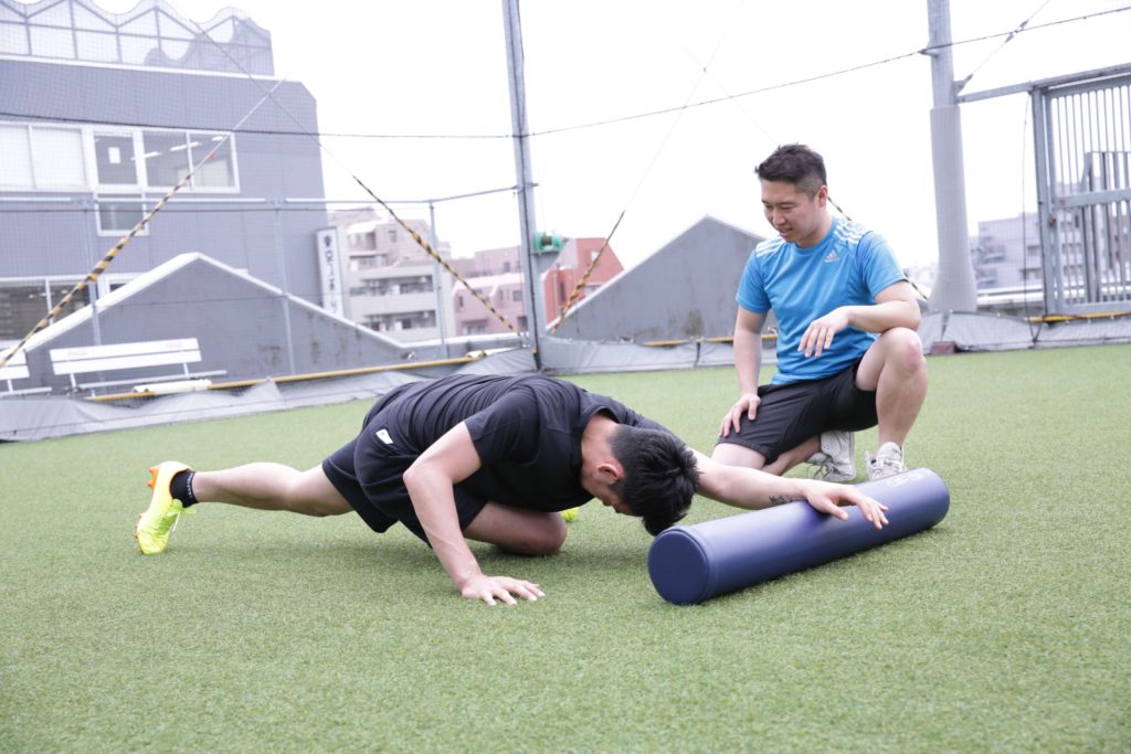 森川稔之が屋外の人工芝で選手のフォームローラーを補助する様子
